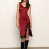 A woman in the Sanctuary Clothing revenge midi satin dress in crimson, featuring a sleeveless design and black lace-trimmed thigh-high slit, paired with knee-high black boots, stands against a plain light background.