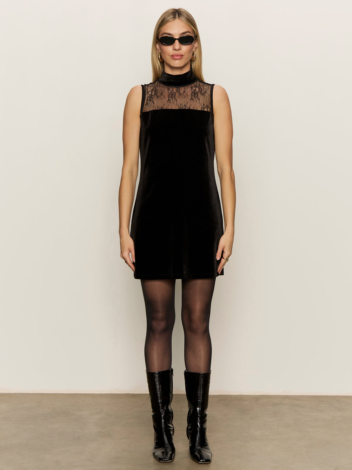 A woman wearing the Sanctuary Clothing party shift dress in black stands against a plain light wall, styled with black tights, knee-high boots, and dark sunglasses.