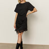 A woman poses against a plain background in the Sanctuary Clothing satin mix tshirt dress black, styled with black knee-high boots and gold hoop earrings. She stands with one hand on her hip and the other relaxed.