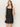 A woman with long brown hair wears the Sanctuary Clothing "almost there linen dress black," a sleeveless style with a square neckline and scalloped hem. She stands against a plain light background, gazing to the side with one hand behind her back.