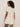 A woman with curly hair models Sanctuary Clothing's For Keeps Bubble Tshirt Dress Collagen, a short light beige dress with a drop waist and bubble hem, as she stands sideways and looks over her shoulder against a plain background.