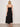 A woman with blonde hair stands with her back to the camera against a light background, wearing Sanctuary Clothing's mira embellished volume dress macro lotus—a black, sleeveless, spaghetti strap midi dress—with brown sandals.