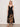 A woman with shoulder-length blonde hair wears the Sanctuary Clothing "mira embellished volume dress macro lotus," a black sleeveless dress featuring thin straps, a deep V-neckline, and bold tan floral prints near the hem. She pairs it with flat sandals.