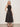 A woman with blonde hair stands against a plain background wearing the Sanctuary Clothing breakaway mix media midi dress in black, featuring a sleeveless fitted top and flared skirt. She pairs it with studded black sandals and has her hand in her pocket.
