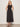 A woman with shoulder-length blonde hair stands by a light wall, wearing Sanctuary Clothing's breakaway mix media midi dress in black and black sandals. The sleeveless dress features a fitted top, button details, and a gathered skirt.