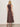 A woman with wavy blonde hair models the Sanctuary Clothing breakaway mix media midi dress in rich soil, facing away from the camera against a plain light beige background. She accessorizes with black sandals and large bracelets.