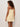 A woman with curly hair wears the Sanctuary Clothing hideaway linen tie strap dress in banana. The light beige, sleeveless, above-the-knee dress has thin shoulder straps tied at the top. She stands facing away, looking over her shoulder against a plain background.