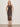 A woman with blonde hair stands against a plain light wall, wearing the Sanctuary Clothing desire more mesh cinch dress rhythmic, a sleeveless fitted black dress with a sheer white-dot overlay, paired with black sandals and her hands behind her back.