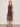 A woman poses against a plain background in the Sanctuary Clothing "desire more mesh cinch dress rhythmic," a sleeveless, sheer, patterned brown midi dress with a keyhole neckline, styled with black sandals and bangle bracelets.