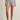 Wearing the Sanctuary Clothing voyage short sail stripe, a person stands against a plain light-colored wall, pairing the navy and white striped shorts with a white top and black flip-flops. Their hands rest by their sides.