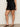 A person wearing Sanctuary Clothing’s almost there linen shorts black extended sizing, featuring a scalloped cut-out hem, and black heeled sandals stands against a plain light-colored wall.