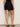 A woman stands facing away, wearing Sanctuary Clothing’s almost there linen shorts black extended sizing with scalloped, laser-cut hems and black heeled sandals against a plain neutral background.