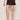 A person wearing Sanctuary Clothing's "wander on short rich soil" stands against a plain light-colored wall, hands in pockets, paired with a striped beige and brown top and brown sandals.