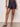 A person wearing Sanctuary Clothing's Summit Short First Sight stands against a plain light background, paired with a patterned sleeveless top, studded sandals, and brown bangles, one leg slightly forward.