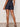 A person standing indoors facing away wears Sanctuary Clothing's summit short first sight denim shorts, a patterned short-sleeve top, and flat studded sandals against a plain light-colored background.