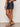 Only the lower half of a person is visible as they stand against a neutral wall, wearing Sanctuary Clothing’s Summit Short First Sight in dark denim, paired with studded black sandals and a brown bangle.
