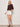 A woman with short blonde hair poses casually against a light background, wearing the Summit Short Chalk by Sanctuary Clothing, styled with a dark brown textured sweater and beige platform sandals.