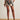 A person stands on a woven rug wearing Sanctuary Clothing's good time linen short lotus in petite sizing, styled with a matching top, brown sandals, and a chunky bracelet, hands in pockets against a plain white wall.