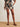 A person stands on a woven rug wearing Sanctuary Clothing's good time linen short lotus in petite sizing, styled with a matching top, brown sandals, and a chunky bracelet, hands in pockets against a plain white wall.