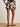 A person stands on a woven rug wearing the Sanctuary Clothing good time linen short in lotus petite sizing, paired with a matching top, tan sandals, and a chunky bracelet, shown from the back against a plain light-colored wall.