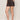 A person stands sideways wearing the Sanctuary Clothing all time linen short rhythmic in a patterned black design, paired with a matching top and brown slide sandals with gold studs. Their left hand is in their pocket, showing a small ankle tattoo.
