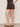 A person stands sideways wearing the Sanctuary Clothing all time linen short rhythmic in a patterned black design, paired with a matching top and brown slide sandals with gold studs. Their left hand is in their pocket, showing a small ankle tattoo.