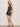 A woman with shoulder-length blonde hair poses relaxed against a light wall, wearing the Sanctuary Clothing all time linen short rhythmic romper with a geometric pattern and brown sandals, her hands in her pockets.