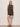 A woman with shoulder-length blonde hair poses against a plain wall wearing the Sanctuary Clothing all time linen short rhythmic—a sleeveless, black patterned romper with shorts and side pockets. She styles it with tan sandals and a necklace, her hands in her pockets.