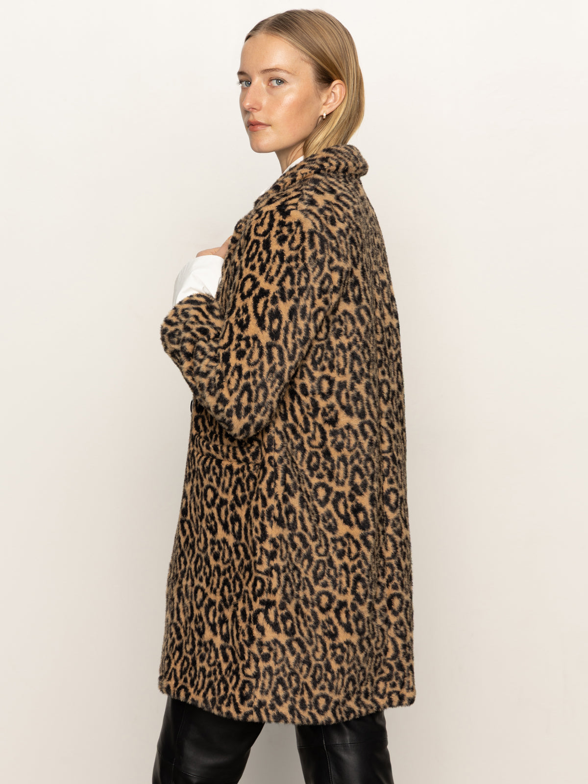 A woman with straight blonde hair models the Sanctuary Clothing hometown jacket lynx, a long leopard-print faux fur coat, paired with black pants. She stands against a plain white background, looking over her shoulder at the camera.