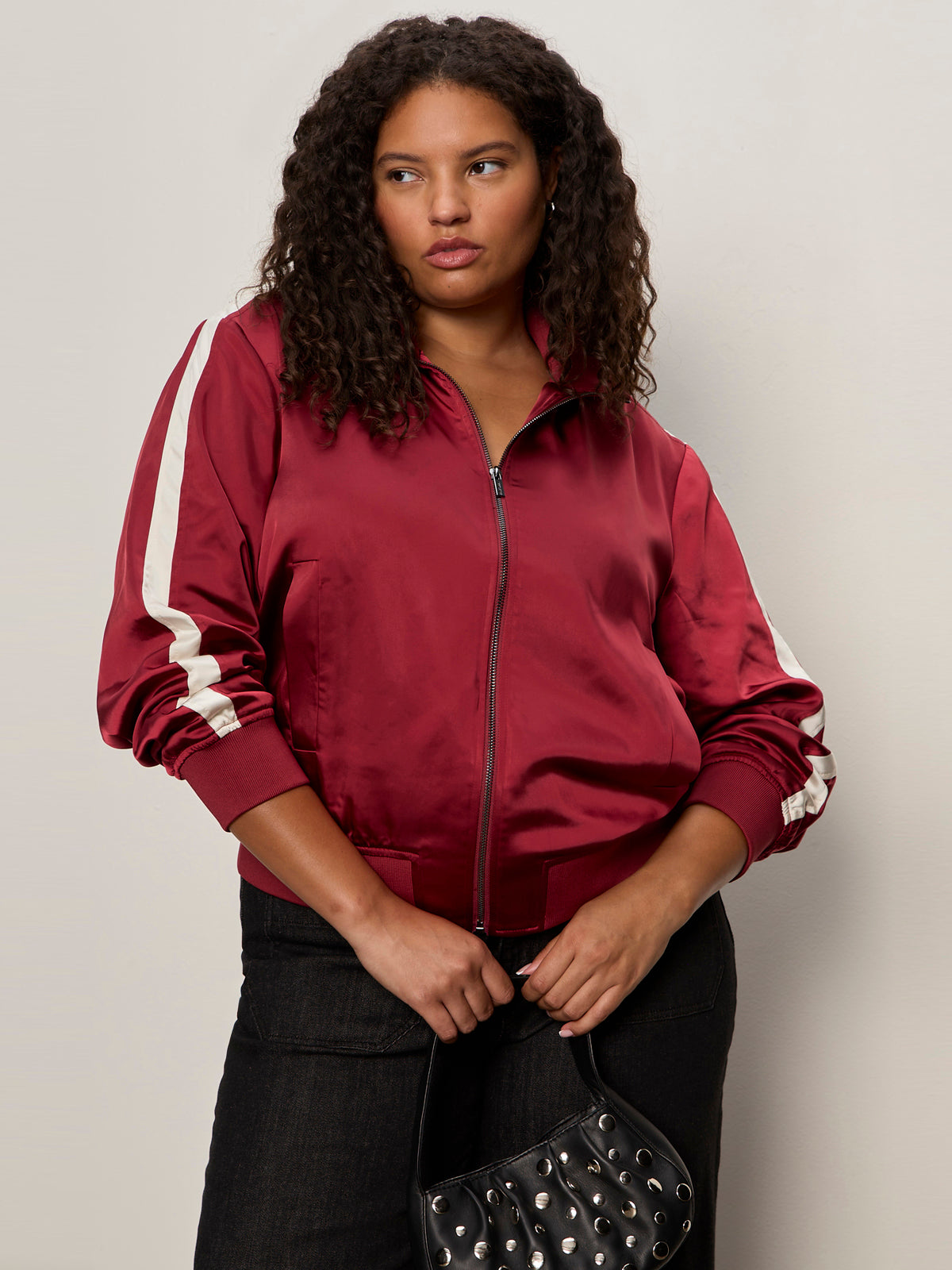A woman with curly hair models the Sanctuary Clothing satin track jacket in crimson (extended sizing), featuring white sleeve stripes. She pairs it with dark pants and a black studded purse, posing against a light background.