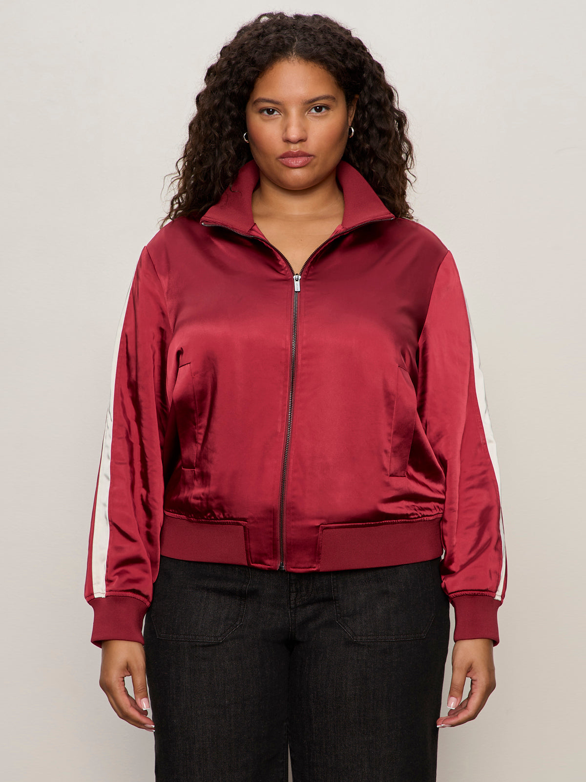 A woman with curly hair wears the Sanctuary Clothing satin track jacket in crimson (extended sizing) with white sleeve stripes and black pants, standing against a plain light background.