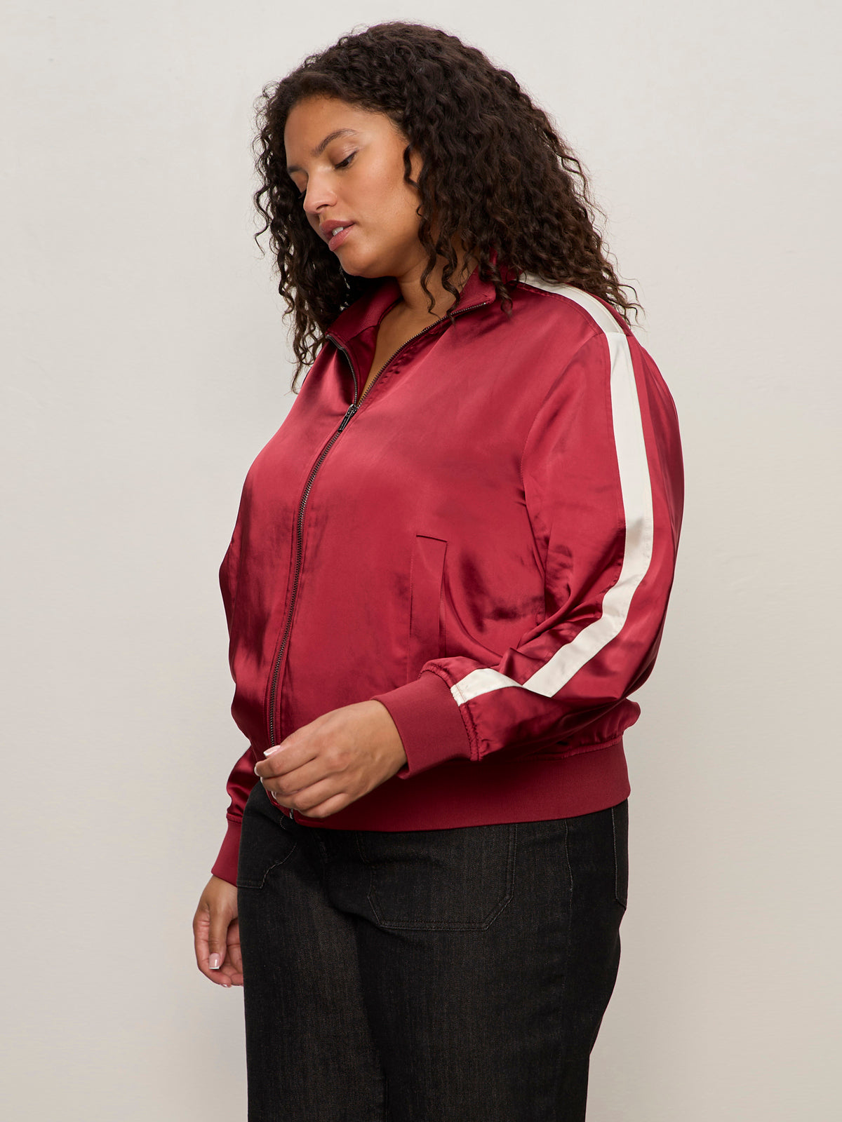 A woman with curly hair wears the Sanctuary Clothing satin track jacket in crimson (extended sizing), featuring white sleeve stripes, and dark pants, standing against a plain light-colored background.