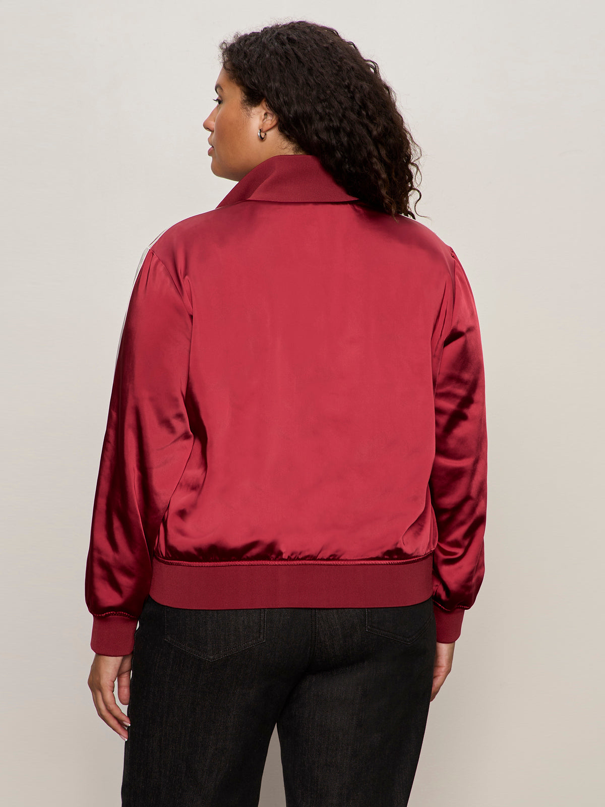 A person with curly hair is seen from the back, wearing the Sanctuary Clothing satin track jacket in crimson (extended sizing) with dark pants, standing against a light beige background.