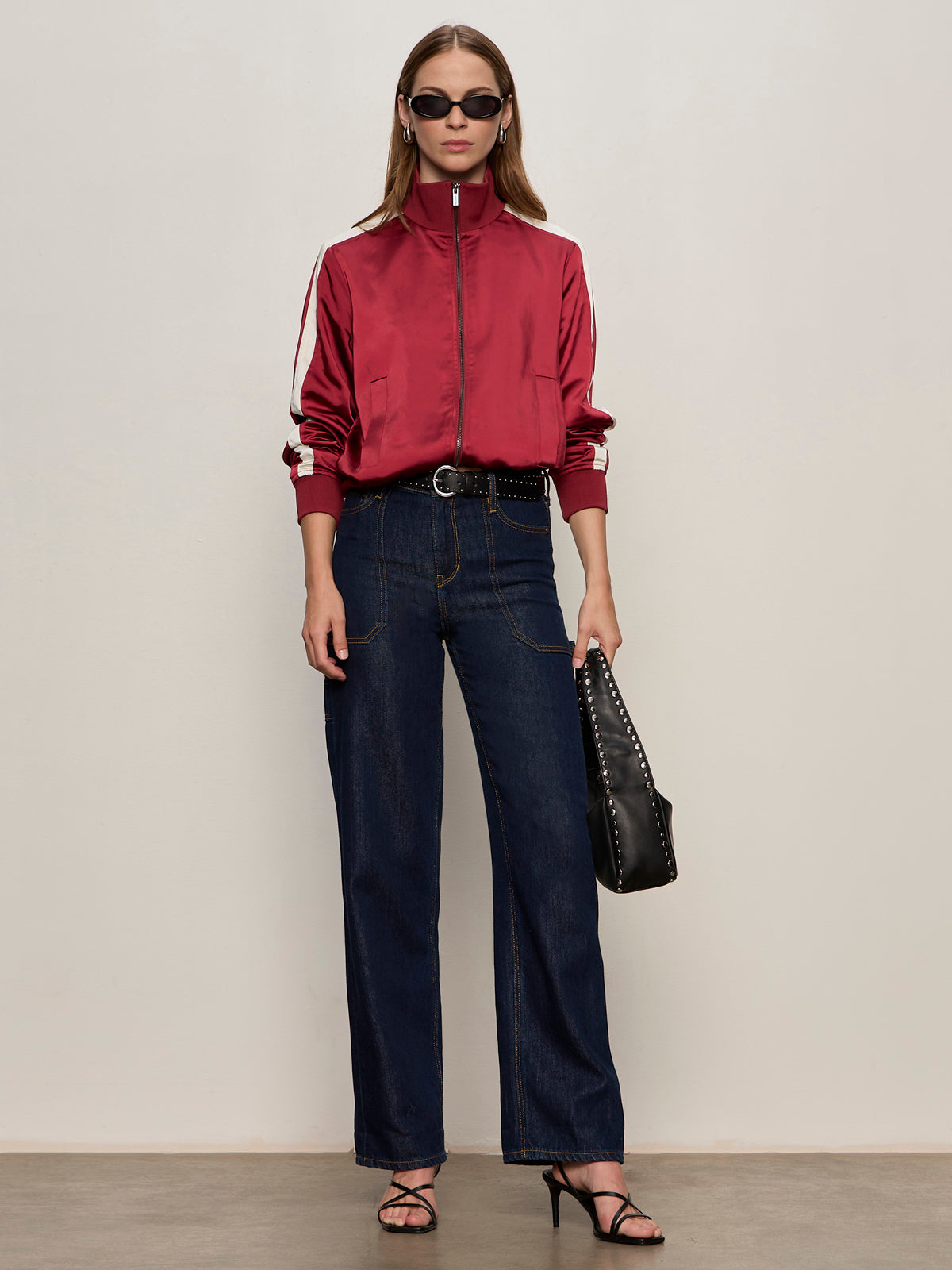 A woman in Sanctuary Clothing's satin track jacket in crimson, sunglasses, wide-leg dark blue jeans, black heeled sandals, and a black studded handbag poses against a plain background.