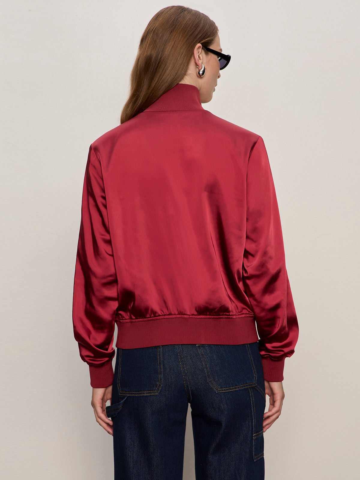 A person with long brown hair and sunglasses faces away from the camera in a Sanctuary Clothing satin track jacket in crimson and dark blue jeans, set against a plain light background.