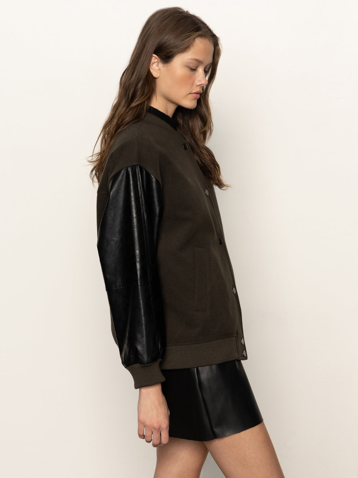 A woman stands in profile against a plain light background, wearing the Sanctuary Clothing club varsity jacket in midnight olive with black leather sleeves and a short black leather skirt.