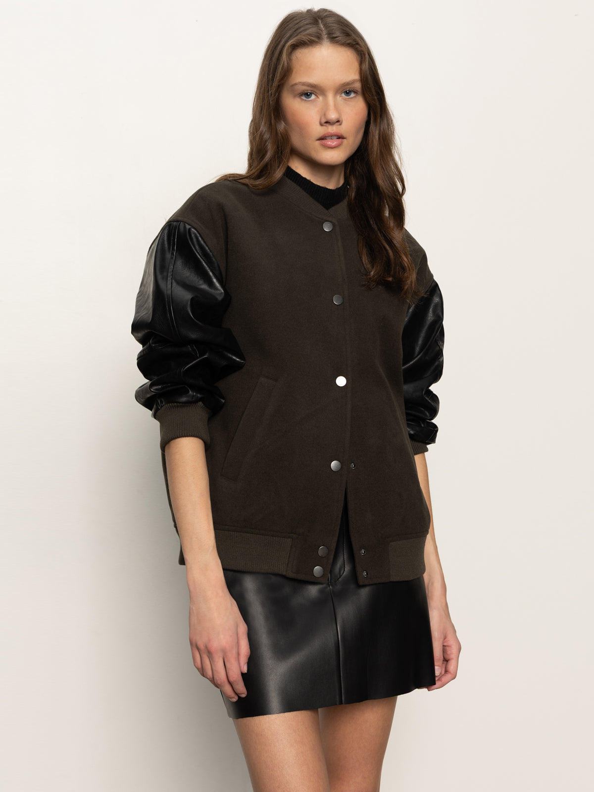 A woman with loose brown hair and a neutral expression stands against a plain background, wearing Sanctuary Clothing's club varsity jacket in midnight olive paired with a black leather mini skirt.
