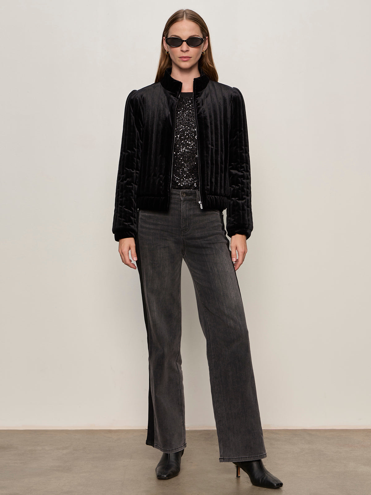A woman stands against a plain wall wearing black sunglasses, a Sanctuary Clothing quilted velvet jacket in black, a sequined top, high-waisted gray wide-leg jeans, and black heeled boots.