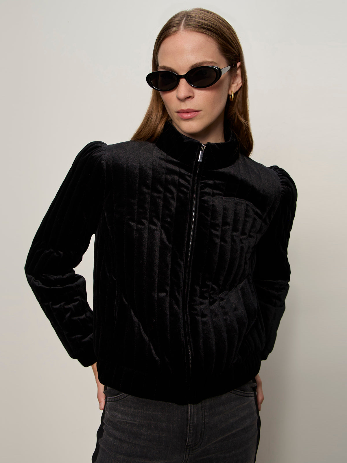 A woman models the Sanctuary Clothing quilted velvet jacket in black, paired with dark sunglasses and gold hoop earrings, standing with her hands in her pockets against a plain background.