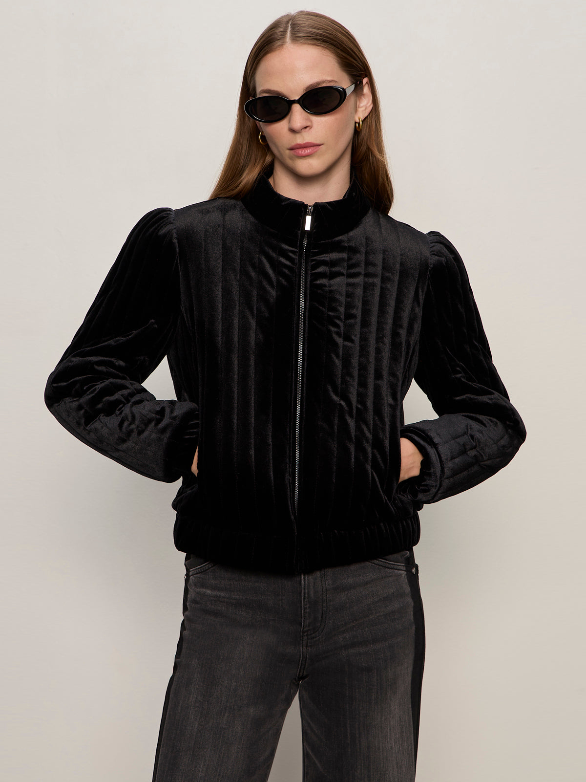 A woman wears Sanctuary Clothing's quilted velvet jacket in black, featuring vertical quilting and puffed sleeves, paired with dark sunglasses and jeans, standing with hands in her pockets against a light background.