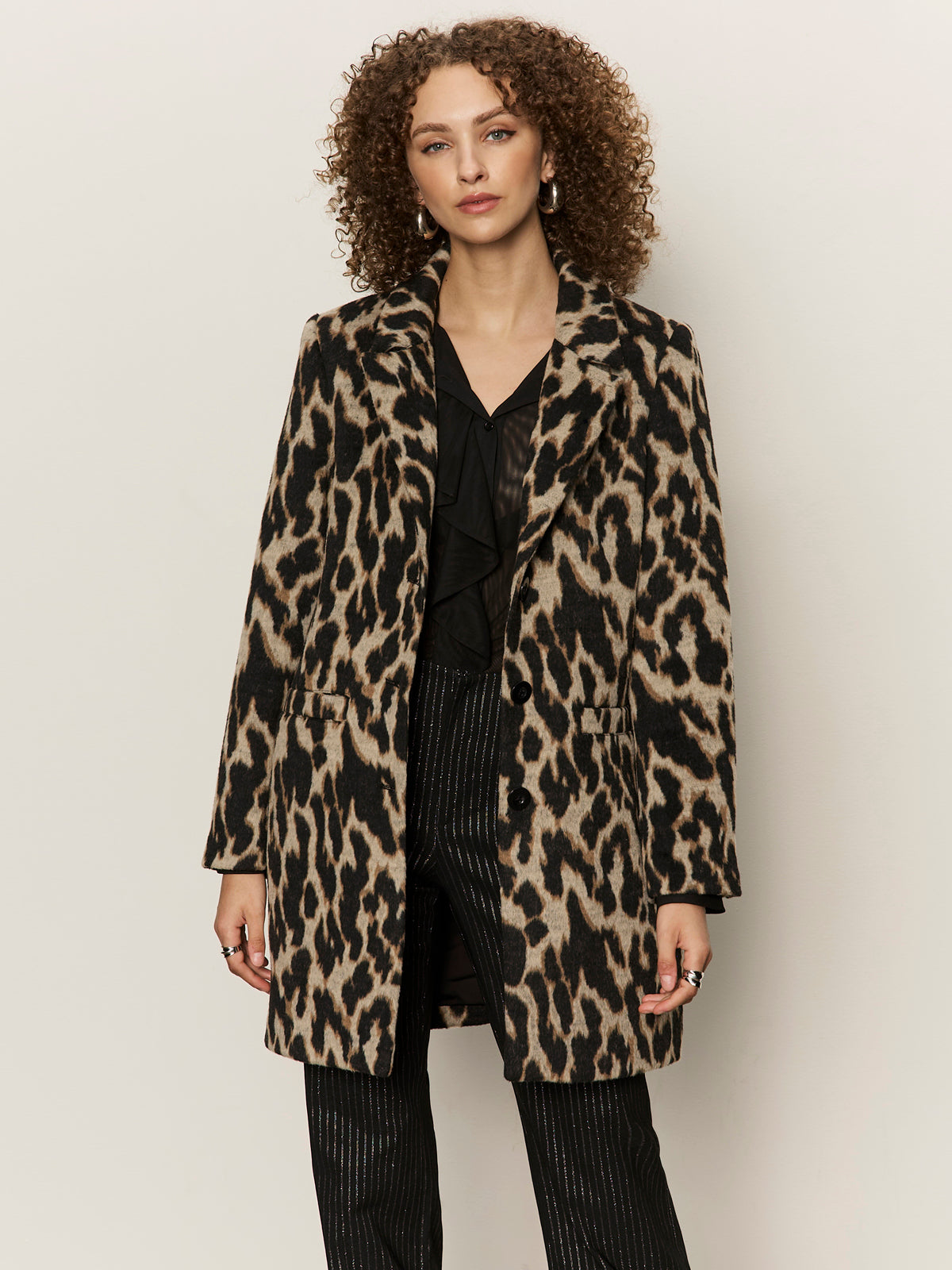 A woman with curly hair models the Sanctuary Clothing downtown jacket leopard mirage over a black blouse and pinstripe pants, standing against a plain light background.