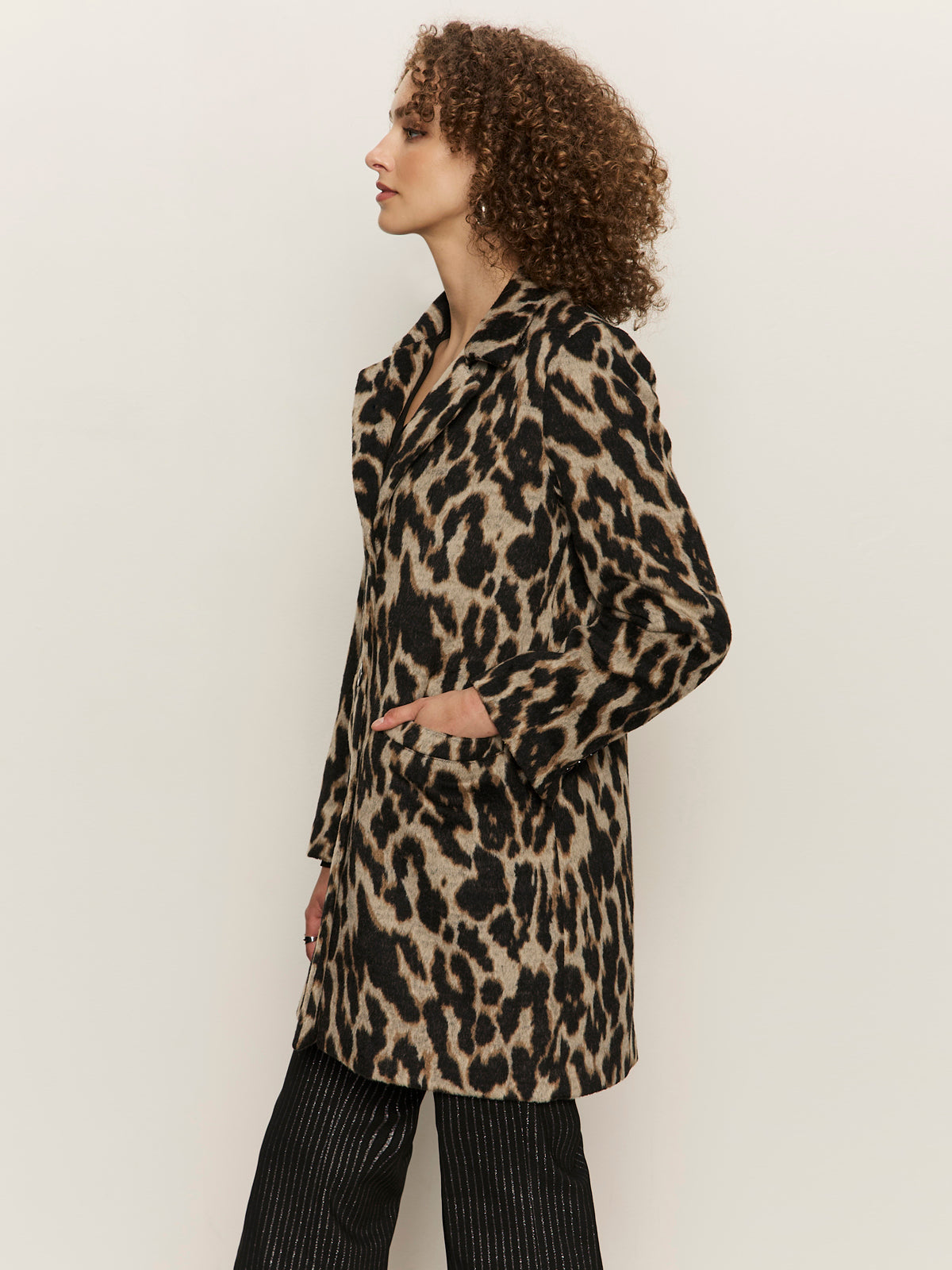 A woman with curly hair stands in profile wearing the Sanctuary Clothing downtown jacket leopard mirage, paired with black pinstripe pants, her hand in the pocket, against a plain light background.