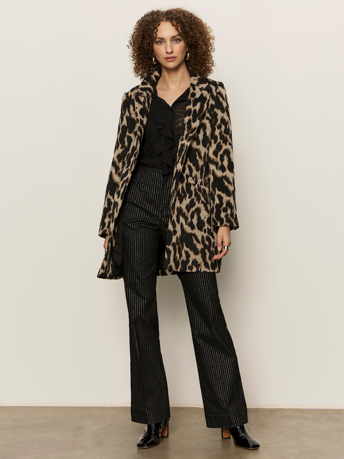 A woman with curly hair stands against a plain background, wearing the Sanctuary Clothing downtown jacket leopard mirage over a black blouse and pinstriped pants, paired with black ankle boots.