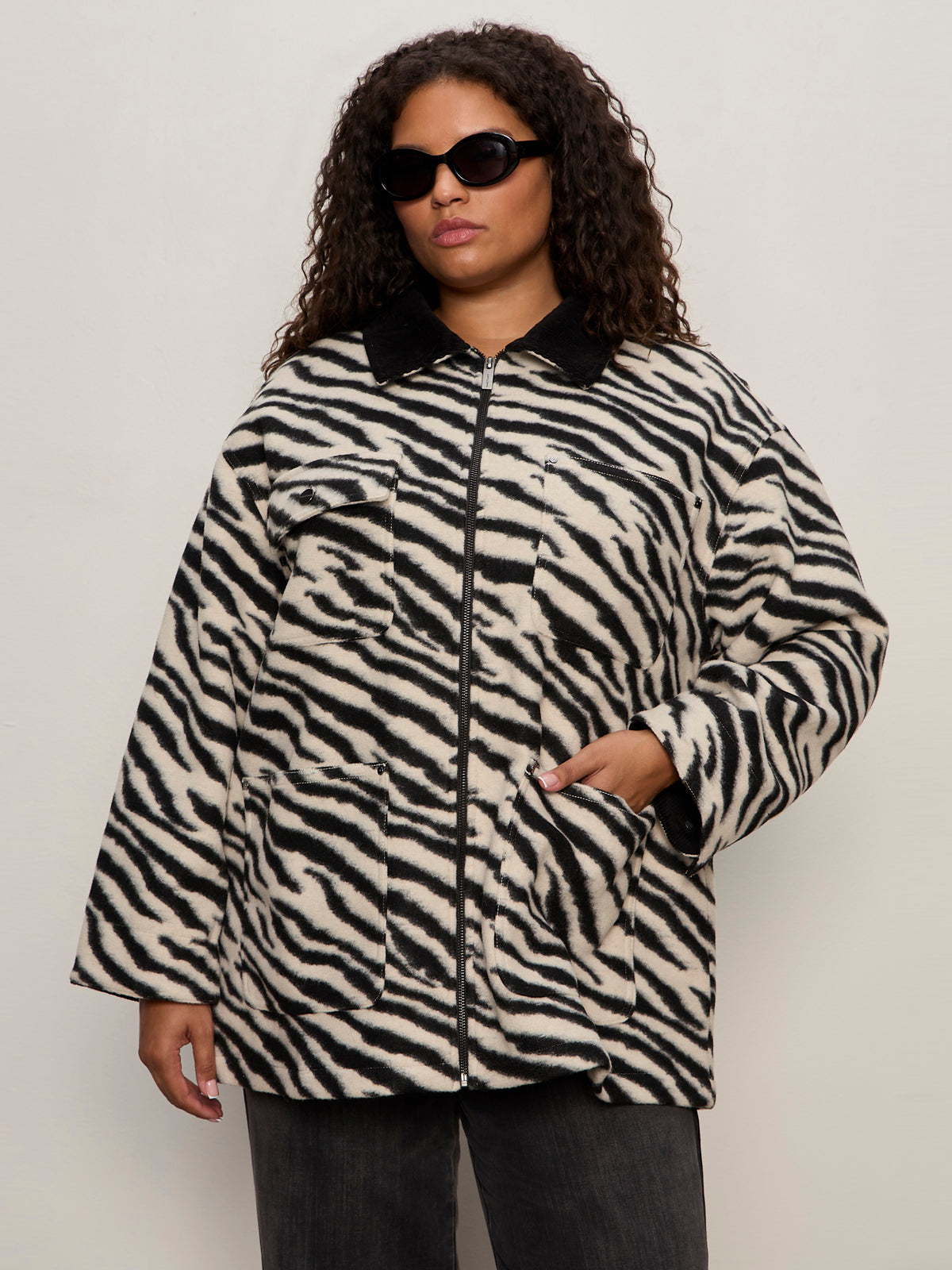 A confident woman with curly hair wears black sunglasses and the Sanctuary Clothing soiree barn jacket in striking zebra extended sizing, standing against a plain light background with one hand in her pocket.