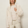 A woman with long blonde hair and minimal makeup stands against a plain light background, wearing Sanctuary Clothing's coco fur jacket in almond cream over a lacy white top and cream pants, holding the jacket collar with one hand.