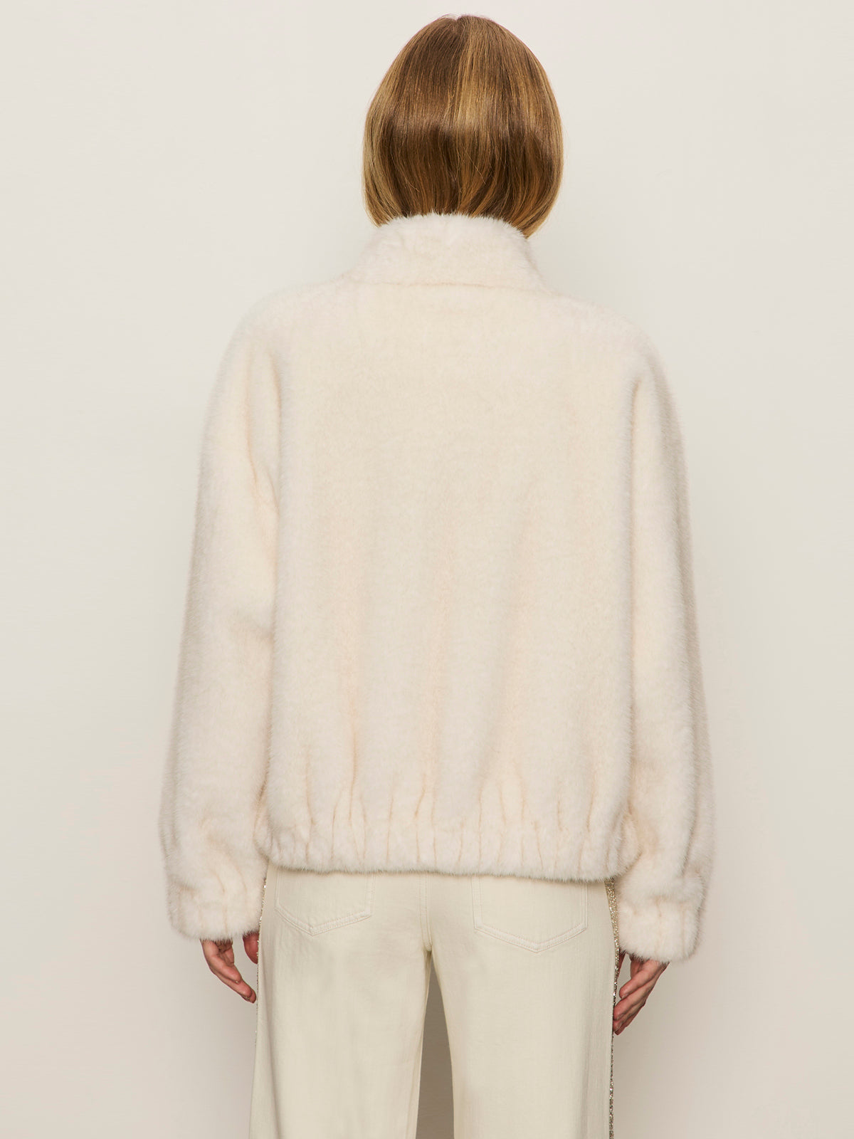 A person with shoulder-length light brown hair, seen from behind, wears the Sanctuary Clothing coco fur jacket in almond cream with light pants against a plain light background.