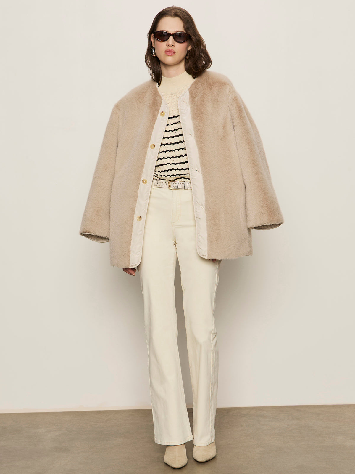 A person wearing Sanctuary Clothing’s reversible fur liner jacket in almond cream, paired with dark sunglasses, a cream and black patterned sweater, cream pants, and beige shoes stands against a plain light-colored wall.