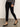 A person wearing Sanctuary Clothing's runway legging black, paired with a gray long-sleeve top and black low-heeled ankle-strap shoes, stands on a smooth floor against a light background, showing only the lower body.