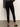 A person stands on a gray floor, shown from the back, wearing Sanctuary Clothing runway legging black, a dark gray long-sleeve top, and black low-heeled ankle-strap shoes.