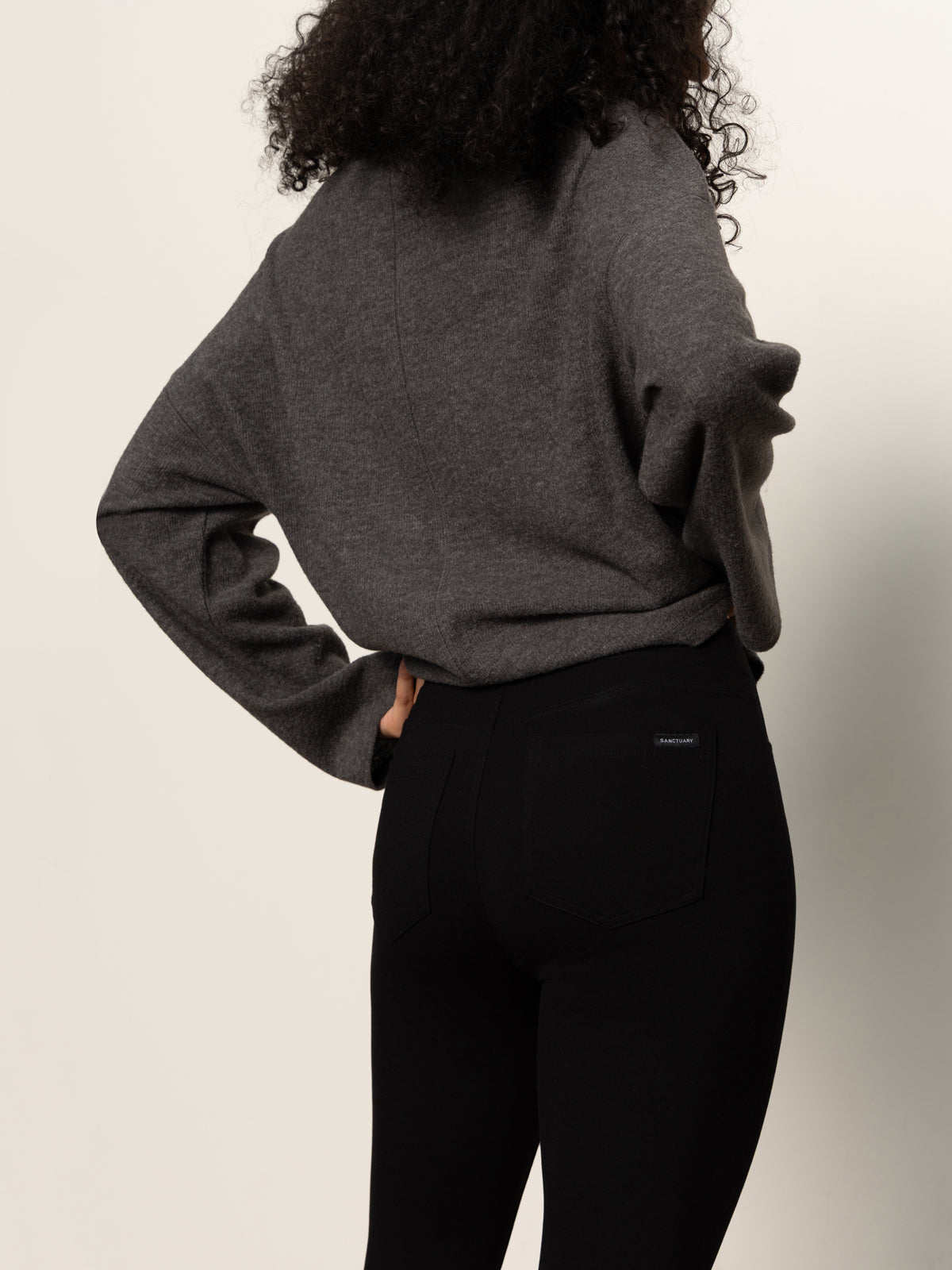 A person with curly hair is shown from the back, wearing a loose gray sweater and Sanctuary Clothing's runway legging black pants against a plain light background.